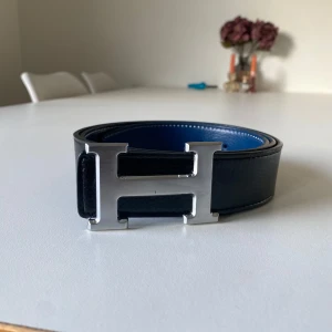 Svart och blått skärp med H-spänne - Säljer ett stilrent skärp i svart och blått med ett stort H-format spänne i silver. Skärpet är vändbart med en sida i svart och den andra i blått, vilket ger två olika looks i ett. Perfekt för både vardag och fest! Skärpet är i mycket bra skick och har en elegant design med vita sömmar.