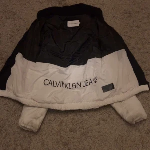  dunjacka från Calvin Klein - Säljer en snygg svart och vit dunjacka från Calvin Klein Jeans. Jackan har en cool färgblockdesign med logga på bröstet och ärmen. Perfekt för kalla dagar med sin varma vaddering. Den har en huva och dragkedja framtill. Passar perfekt för både vardag och festliga tillfällen!