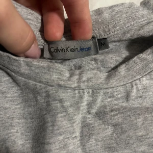 Grå t-shirt från Calvin Klein - Säljer en stilren grå t-shirt från Calvin Klein i bomull. Den har korta ärmar och texten 'Calvin' tryckt på framsidan. Perfekt för en avslappnad look eller vardagsbruk. Passar bra till jeans eller shorts! Knappast andvänd!