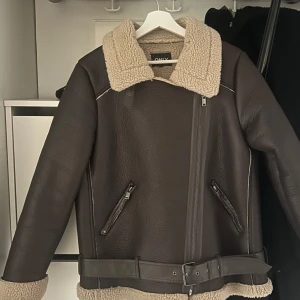 Brun jacka med teddyfoder från ONLY - Säljer en snygg brun jacka från ONLY i storlek M. Den har ett mysigt teddyfoder och en cool bikerstil med dragkedja och bälte i midjan. Perfekt för höst och vinter! 🧥