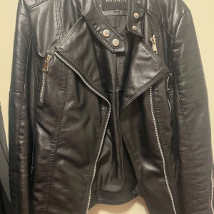 Svart skinnjacka från Missay - Säljer en snygg svart skinnjacka från Missani i storlek M. Jackan har en cool bikerstil med dragkedjor och knappar som detaljer. Perfekt för höst och vår, och ger en edgy touch till vilken outfit som helst. Passar både till vardags och fest!