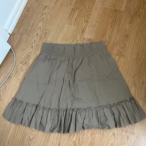 Beige kjol med volang - Säljer en superfin beige kjol med volangdetaljer längst ner. Den har en elastisk midja för en bekväm passform. Perfekt för sommardagar eller en avslappnad vårlook. Kjolen är i bra skick och passar till både vardag och fest.