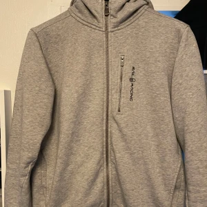 Grå hoodie från Sail Racing - Säljer en snygg grå hoodie från Sail Racing i storlek M. Den har en dragkedja framtill och en praktisk ficka med dragkedja på bröstet. Perfekt för kyliga dagar och passar till både vardag och träning. Huvan har en stilren design med en liten logga på baksidan.