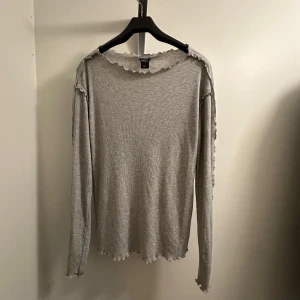 Grå ribbad tröja med volangdetaljer - En superfin grå ribbad tröja från Lindex i storlek L. Den har långa ärmar och söta volangdetaljer vid halsen och ärmsluten. Helt oanvänd!