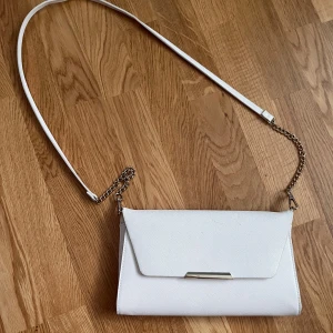 Vit kuvertväska från H&M - Säljer en stilren vit kuvertväska från H&M. Den har en elegant kedjedetalj på axelremmen och stängs med en tryckknapp. Perfekt för festliga tillfällen eller en utekväll. Väskan är i bra skick, men lite smutsig. Har en praktisk innerficka med dragkedja🤍