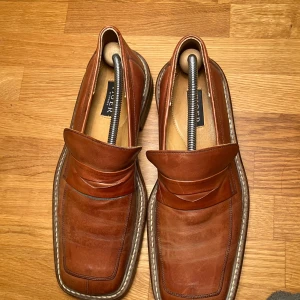 Bruna loafers från Tiger - Säljer ett par snygga bruna loafers från Tiger i bra skick. De har en stilren design med en fyrkantig tå och är perfekta för både vardag och fest. Skorna har en robust sula och är gjorda i skinn, vilket ger dem en klassisk look. Perfekta för höst och vår!