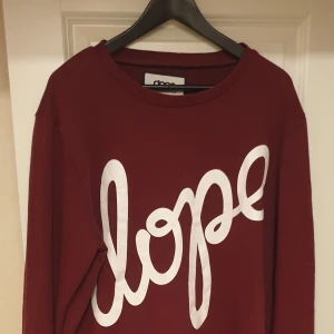 Vinröd tröja från Dope - Säljer en snygg vinröd tröja från Dope med stort tryck 'dope' i vitt på framsidan. Tröjan är i bra skick och perfekt för en avslappnad stil. Den har en normal passform och är gjord i ett mjukt material som är skönt att ha på sig. Perfekt för höst och vår!