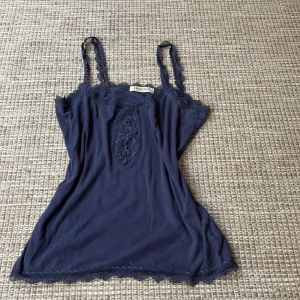 Mörkblå linne från Indigo Collection - Säljer ett mörkblått linne från Indigo Collection. Det har spetsdetaljer vid halsringningen och axelbanden, samt en liten knapp framtill. Perfekt för sommardagar eller som ett lager under en tröja.