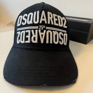 Snygg svart keps från Dsquared2 med vit broderad text på framsidan som firar 25-årsjubileet. Justerbar rem baktill för perfekt passform. Perfekt för både vardag och fest!