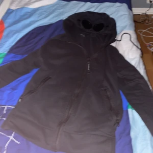 Svart hoodie med huva från C.P. Company - Säljer en svart jacka från C.P. Company i 100% bomull. Den har en cool huva med inbyggda glasögon och dragkedja framtill. Perfekt för höst och vår, och den är i bra skick. Passar både till vardags och lite mer avslappnade tillfällen.