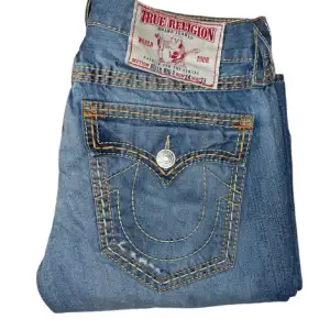 Säljer ett par snygga blå jeans från True Religion. De har en klassisk femficksdesign med coola kontrastsömmar och en unik bakficka med knapp. Perfekta för en avslappnad stil med en touch av lyx. Passar både till vardags och fest!