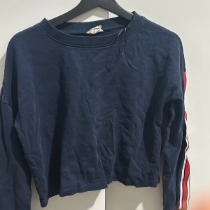 Mörkblå croppad tröja från H&M - Säljer en mörkblå croppad tröja från H&M i ekologisk bomull. Tröjan har långa ärmar med ränder i rött och vitt längs sidorna. Perfekt för en avslappnad stil eller sportig look. Passar bra till både jeans och joggers!
