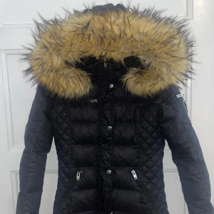 Svart vinterjacka från Rockandblue - Säljer en snygg svart vinterjacka från Rockandblue i storlek 34. Jackan har en fluffig fuskpäls på huvan och quiltad design. Perfekt för kalla vinterdagar med dragkedja och knappar för extra värme. Den är i mycket bra skick och redo för nya äventyr! 🖤