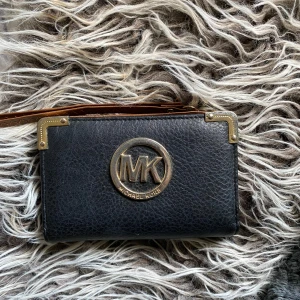 Svart plånbok från Michael Kors - Snygg svart plånbok från Michael Kors i skinn med guldiga detaljer och logga framtill. Insidan är brun med flera fack och dragkedja för mynt. Perfekt för att hålla ordning på kort och kontanter. Stilren och praktisk för vardagsbruk.