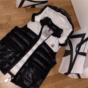 Moncler maya väst - Säljer en snygg svart och vit dunväst från Moncler i mycket bra skick. Västen har en dragkedja framtill och en stor logga på ryggen. Perfekt för höst och vinter, håller dig varm och stilren. Nypris runt 7.9 🤙🏼bra skick 