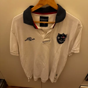 Vit pikétröja från Sebago - Säljer en klassisk vit pikétröja från Sebago i storlek XL. Tröjan har en mörkblå krage och broderad logga på bröstet. Perfekt för en avslappnad men stilren look. Passar både till vardags och lite finare tillfällen. Skön och luftig för varmare dagar.