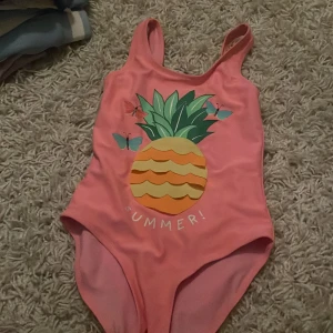 Rosa baddräkt med ananastryck från H&M - Superfin rosa baddräkt från H&M med ett coolt ananastryck och texten 'Summer!' på framsidan. Perfekt för sommarens badäventyr! Den har breda axelband och är gjord i ett mjukt och bekvämt material. Passar barn i storlek 110/116. 🌞🍍