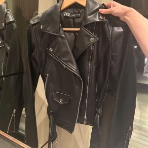 Svart skinnjacka från Zara - Säljer en snygg svart skinnjacka från Zara i mycket bra skick. Den har en cool bikerstil med dragkedja och knappar, samt ett bälte i midjan. Perfekt för höst och vår, och funkar lika bra till fest som till vardags. Jackan har långa ärmar och en klassisk krage.