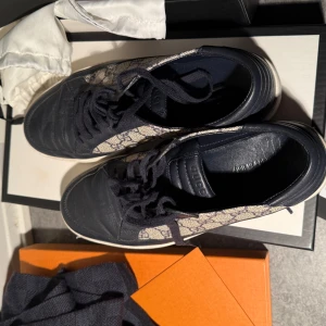 Gucci sneakers med monogram - Säljer ett par snygga Gucci sneakers i mycket bra skick. Skorna har ett klassiskt monogrammönster i beige och blått med mörkblå läderdetaljer. Perfekta för både vardag och fest! Storlek är inte specificerad, men de ser ut att vara i herrstorlek. Skorna har snörning och en vit sula.