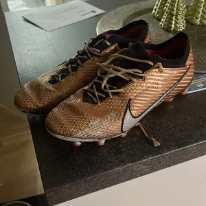 Guldiga fotbollsskor från Nike - Säljer ett par snygga guldiga Nike fotbollsskor i bra skick. De har en cool design med svarta detaljer och snörning. Perfekta för fotbollsspel på gräs med sina robusta dobbar. Skostorlek är herr 44, och de är bekväma för intensiva matcher.