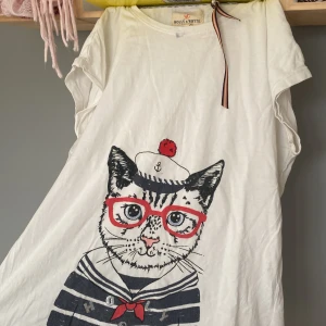 Vit t-shirt med kattmotiv från Holly & White - Säljer en superfin vit t-shirt från Holly & White med ett gulligt kattmotiv. Katten har en sjömanshatt och röda glasögon, vilket ger en lekfull touch. Perfekt för en avslappnad dag eller sommarhäng. T-shirten har korta ärmar och är i ett mjukt bomullsmaterial.