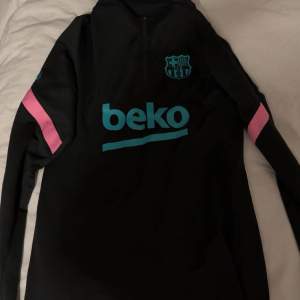 Säljer en svart långärmad träningströja från Nike med Dri-FIT-teknologi. Tröjan har turkosa och rosa detaljer samt en halv dragkedja framtill. Perfekt för fotbollsträning eller andra sportaktiviteter. Bekväm och stilren design!