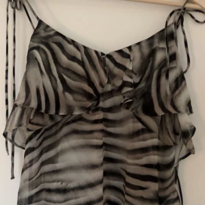 Zebramönstrad topp från Zara - Snygg zebramönstrad klänning från Zara i svart och grått. Klänningen har tunna axelband och volangdetaljer som ger en trendig look. Perfekt för en sommarfest eller en utekväll. Ny med tag kvar! Nypris:379kr
