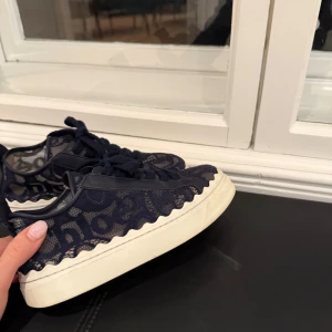 Mörkblå sneakers från Chloé - Snygga mörkblå sneakers från Chloé med unikt spetsmönster och vågig kant. De har en vit sula och snörning framtill. Perfekta för en stilren och trendig look. Märkesloggan syns tydligt på baksidan av skorna. Kartong medföljer! 