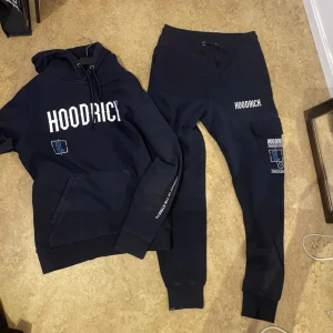 Svart hoodie och joggers från Hoodrich - Säljer ett set med svart hoodie och matchande joggers från Hoodrich. Hoodien har en stor logga på bröstet och ryggen, och byxorna har loggor på benet. Perfekt för en avslappnad stil eller streetwear-look. Setet är i mycket bra skick och redo för nya äventyr!