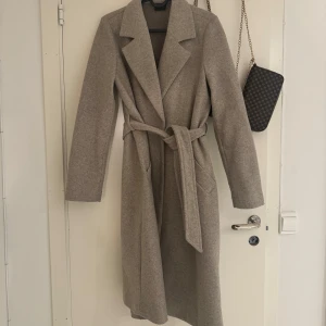 Beige kappa med bälte - Säljer en beige kappa i mycket bra skick. Storlek XS. Den har en klassisk design med långa ärmar och ett bälte i midjan för en snygg passform. 