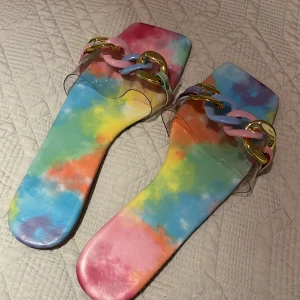 Färgglada sandaler med kedjedetalj - Supercoola sandaler i regnbågens alla färger! De har en transparent rem med en stor, färgglad kedja som detalj. Perfekta för sommaren när du vill sticka ut lite extra. De är platta och bekväma, så du kan ha dem på dig hela dagen. Passar perfekt till en avslappnad sommarlook eller en dag på stranden.