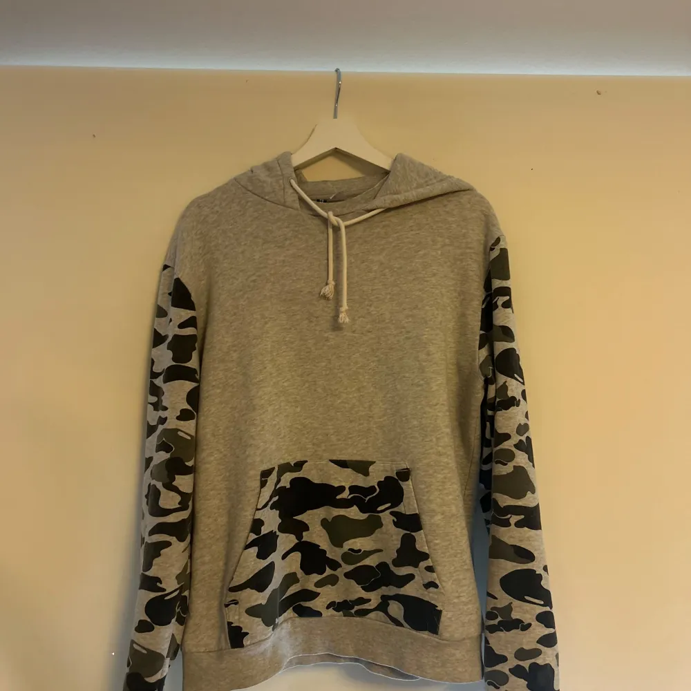 Säljer en snygg grå hoodie med coola camouflageärmar och ficka. Perfekt för en avslappnad stil och passar bra till både höst och vår. Den har en justerbar huva med snören och är i mycket bra skick. Perfekt för dig som gillar streetwear!. Tröjor & Koftor.