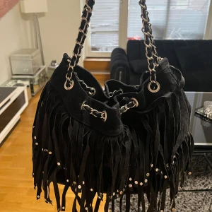 Svart axelväska med fransar - Snygg svart axelväska i mocka med coola fransar och silverdetaljer. Väskan har en kedja som axelrem samt ett längre band och är i mycket bra skick. Pris kan diskuteras 🤎