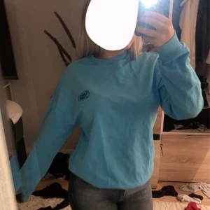 Blå långärmad tröja med broderad logga - Säljer en snygg blå långärmad tröja med en broderad logga på bröstet. Perfekt för en casual look och superbekväm att ha på sig. Tröjan är i bra skick och passar perfekt till jeans eller leggings. Perfekt för höst och vår!