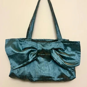 Blå axelväska med rosett - Snygg blå axelväska i glansigt material med en stor dekorativ rosett på framsidan. Väskan har en rymlig insida med foder. Passar både till vardags och festliga tillfällen. Mått: 28x16cm