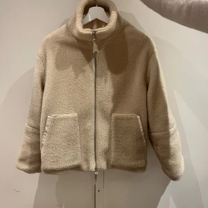 Beige teddyjacka med dragkedja - Mysig beige teddyjacka i mycket bra skick. Den har en dragkedja framtill och två stora fickor. Perfekt för höst och vinter när du vill hålla dig varm och stilren. Jackan har en normal passform och långa ärmar.