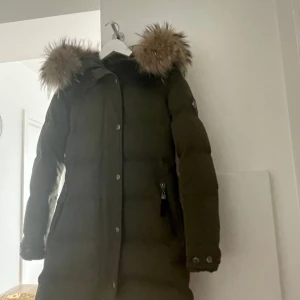 Olivgrön dunjacka med pälsdetalj - Säljer en olivgrön dunjacka med stor, fluffig pälsdetalj på huvan. Jackan har långa ärmar och knappar längs framsidan. Perfekt för kalla vinterdagar och håller dig varm och stilren. Passar både till vardags och lite finare tillfällen.