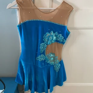 Blå konståkningsklänning - Säljer en superfin blå konståkningsklänning med blommiga paljettdetaljer. Klänningen har en ärmlös design och en mesh-överdel med strass. Perfekt för tävlingar eller träning på isen! Endast använd 1 gång 💙
