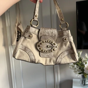 Vintage Guldig axelväska från Guess - Snygg guldig axelväska i syntetmaterial med coola nitar och ett stort spänne framtill. Den är vintage från guess. Har inte koll på nypriset.
