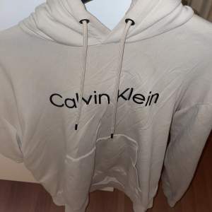 Säljer en snygg beige hoodie från Calvin Klein i storlek M. Den har en klassisk design med loggan tryckt på framsidan och en bekväm passform. Perfekt för både vardag och chill. Huvan har dragsko och materialet känns mjukt och skönt. Passa på att fynda denna stilrena tröja!