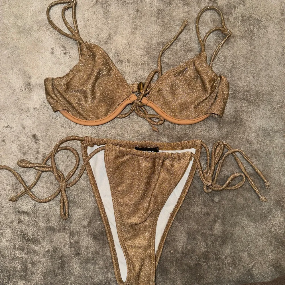 Snygg glittrig bikini i guld med knytband på sidorna och axelbanden. Bikinin har en stilren design med en liten detalj framtill på toppen. Materialet är mjukt och bekvämt, vilket gör den idealisk för både bad och solning. Passar perfekt för dig som vill glänsa lite extra i solen! Från Boohoo och storlek xs . Övrigt.