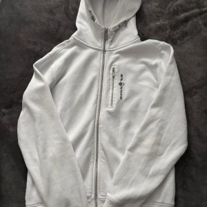 Vit huvtröja från Sail Racing - Säljer en vit huvtröja från Sail Racing i bra skick. Den har en dragkedja framtill och en praktisk bröstficka med dragkedja. Perfekt för kyliga dagar och passar till både vardag och träning. Huvan är justerbar med snören.