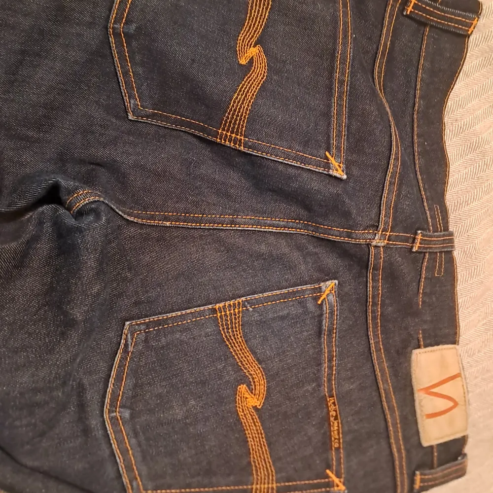 Säljer ett par snygga mörkblå jeans från Nudie Jeans i bra skick. De har en klassisk femficksdesign med orange sömmar och en snygg logga på bakfickan. Perfekta för både vardag och fest! Passar till allt och är superbekväma. De är som nya och använda 2 gånger de är i storleken W 31, L32. Jeans & Byxor.