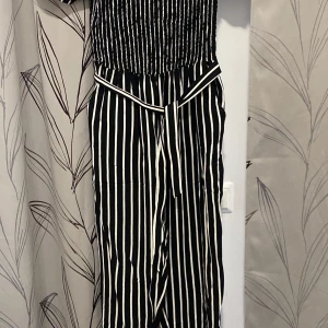 Svartvit randig långklänning - Säljer en superfin svartvit randig overall med knytband i midjan. Den har en stilren design med vertikala ränder som ger en smickrande siluett. Perfekt för både vardag och fest!