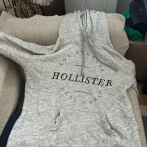 Grå hoodie från Hollister - Säljer en supermjuk och bekväm grå hoodie från Hollister. Den har en klassisk design med 'Los Angeles West Coast' tryckt på framsidan. Perfekt för en avslappnad stil och passar bra till både jeans och mjukisbyxor. Den har en känguruficka och en justerbar huva. Perfekt för kyliga dagar!