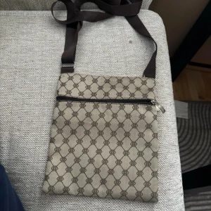 Beige axelväska med mönster - Säljer en snygg beige axelväska med ett klassiskt mönster. Väskan har en praktisk dragkedja och en justerbar axelrem. Perfekt för vardagsbruk eller när du vill ha med dig det viktigaste på stan.