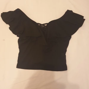 Topp - Svart topp med spetsdetaljer i ryggen och volanger på axlarna. Toppen är kort och har en avslappnad passform. Säljer för 40