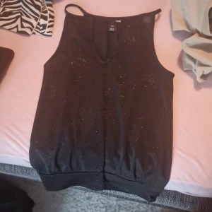 Svart glittrig ärmlös topp från H&M - Säljer en snygg svart glittrig topp från H&M i storlek XS. Den är ärmlös och perfekt för festliga tillfällen. Toppen har en bekväm passform och är gjord i ett lätt material som glittrar fint i ljuset. Perfekt för att piffa upp vilken outfit som helst!
