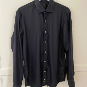 Svart skjorta från Dahlin - Säljer en stilren svart skjorta från Dahlin i storlek M. Skjortan har långa ärmar och knappar framtill. Perfekt för både vardag och festliga tillfällen. Den är i nyskick och har en snygg detalj med märkets logga vid nederkanten.