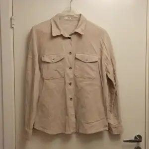 Säljer en snygg beige overshirt/skjorta från MNG Casual. Den har en avslappnad stil med knappar framtill och två bröstfickor. Materialet känns mjukt och bekvämt, och den har långa ärmar med knappar. Det saknas 2 knappar, en högst upp & en längst ner.